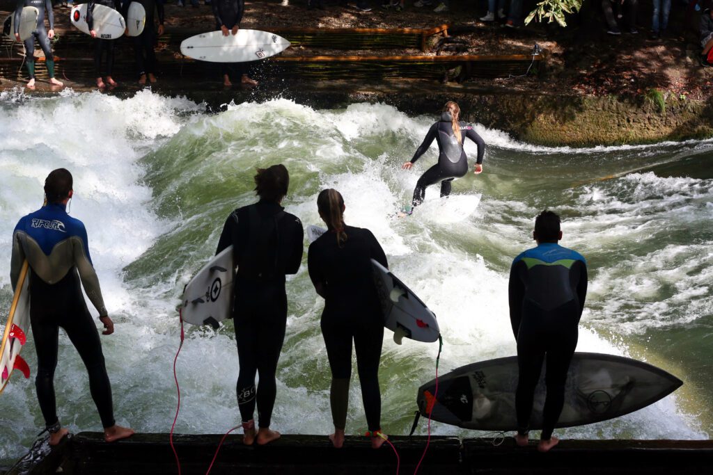 One surfer is rising the wave, while several surfers on both sides of the stream look on and wait for their turn