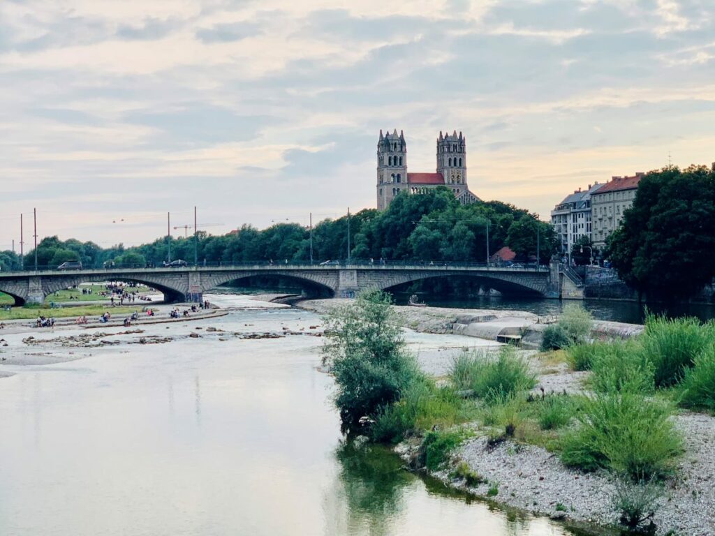 Isar river in Munich, Germany, with view of bridge and church in the background and people sitting on the banks of the river